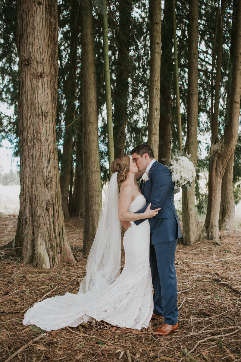 real bride wearing a floor length veil holding a white bouquet and kissing her groom in the forest