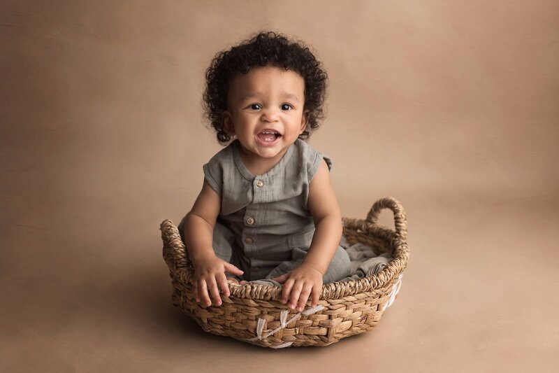 A toddler in a grey onesie sits in a woven basket in a Lafayette Baby Milestone Photographer studio