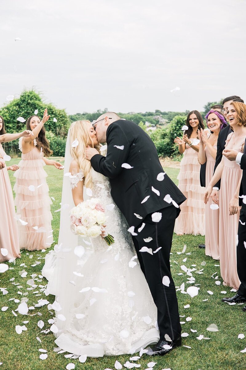 Bride and Groom kissing. Flower petals float in air around them with smiling bridal party faces surrounding them.