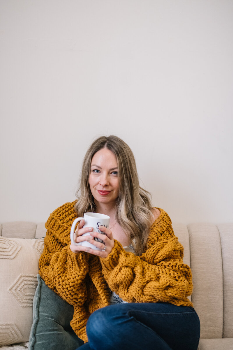 Sammy in a yellow cardigan sitting on a couch holding a mug