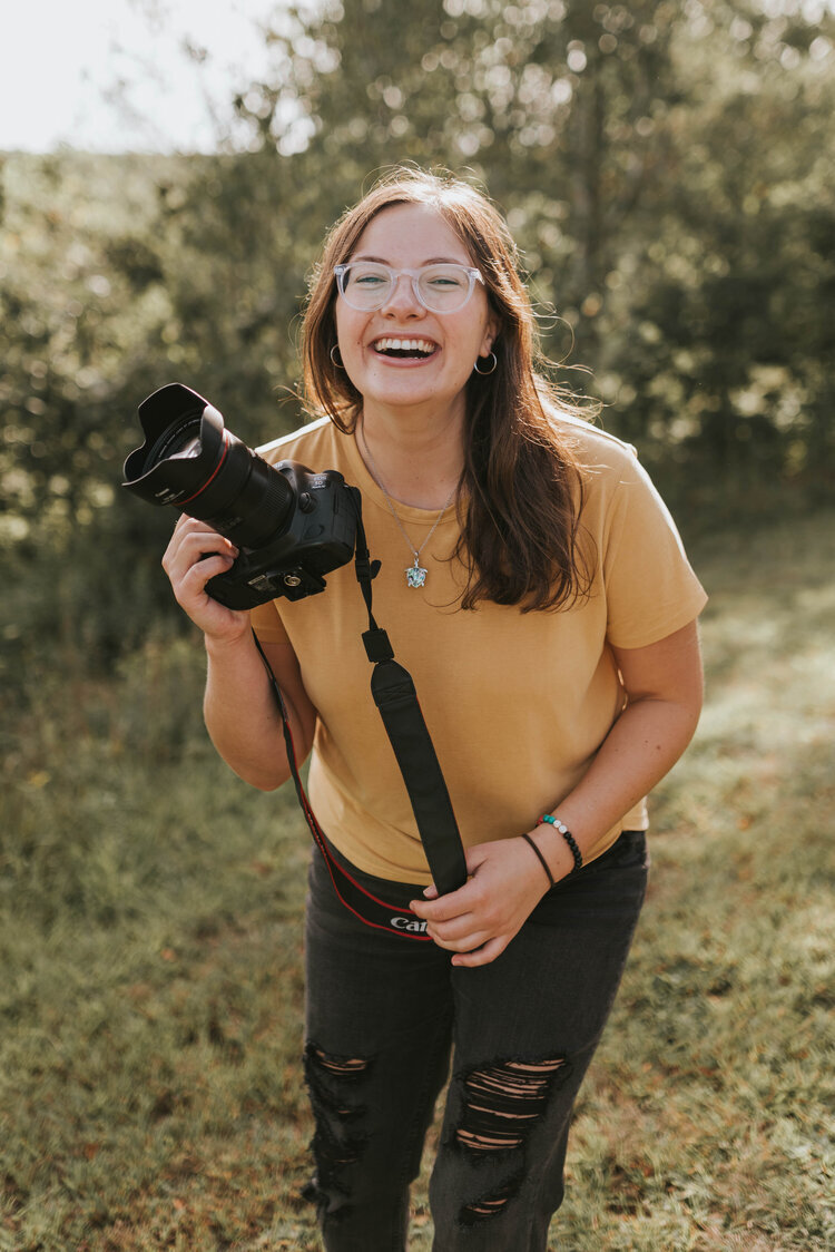 woman smiling while holding camera and wearing a mustard shirt and jeans