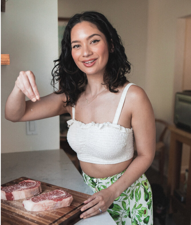 A smiling woman with shoulder-length dark curly hair, standing in a kitchen setting. She is wearing a white, ruffled crop top and green leaf-patterned bottoms. The woman is seasoning a piece of raw steak on a wooden cutting board, which suggests she may be preparing to cook. Her pose and the domestic environment suggest a relaxed and cheerful cooking atmosphere.