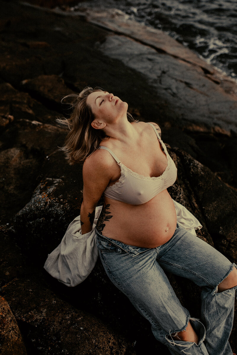 en gravid kvinna sitter stolt på klippor med huvudet kastat bakåt iklädd jeans och en skjorta som hänger över axlarna