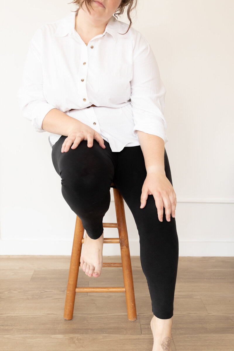 emily of emmy & ollie photographer sits on a stool at StudioStudio in Ann Arbor michigan for branding session wearing white button up and black leggings