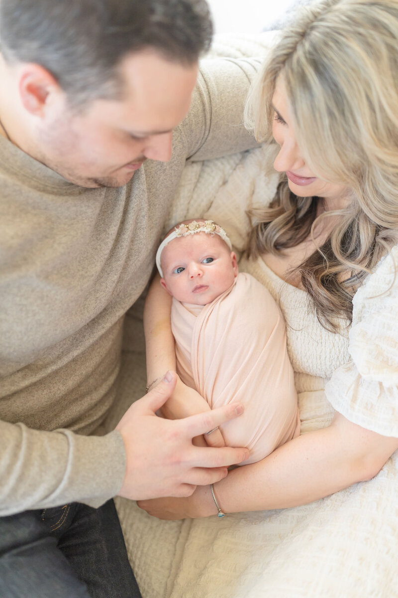 Mom and Dad hold their newborn together, gazing lovingly at their baby. Their faces reflect warmth and tenderness, capturing a heartfelt moment during the newborn session. Soft, natural light enhances the intimate and cozy atmosphere