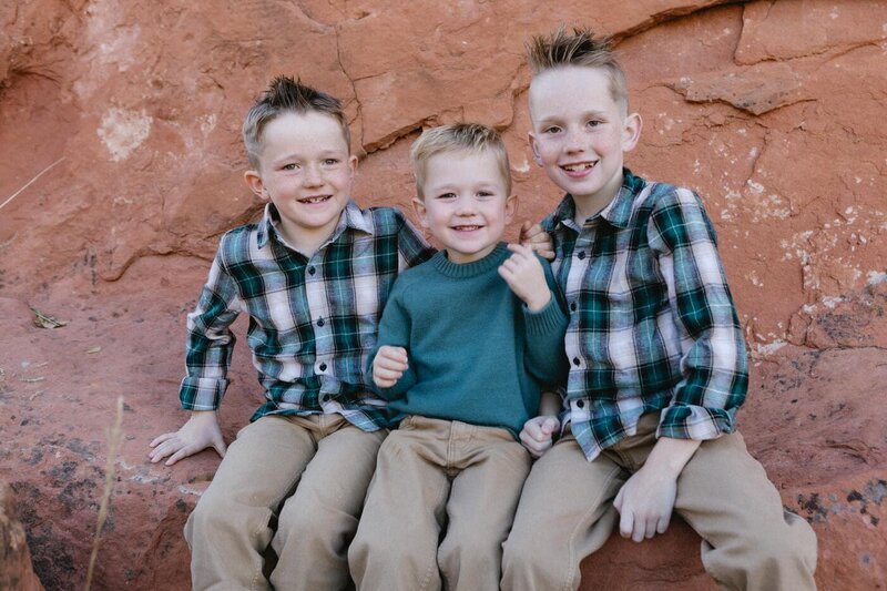 Three school-aged brothers sit atop a red rock and smile at the camera.