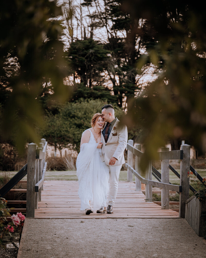 Bride and groom walking along a bridge surrounded by foliage laughing