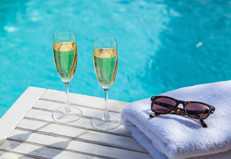 Two glasses of champagne on a poolside table next to a folded towel and sunglasses.