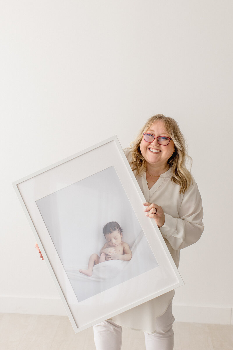 photographer standing in white studio holding large framed portrait of a cute newborn