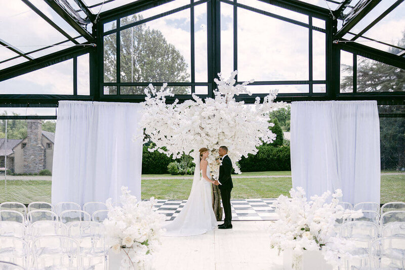 A splendid wedding ceremony unfolds in Glenstone gardens, Virginia, within a stunning glass greenhouse embellished with exquisite floral arrangements and refined decorations.
