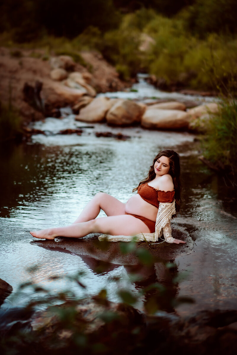 Pregnant mother sitting in stream in estes park colorado wearing brown two piece and ivory lace shawl.