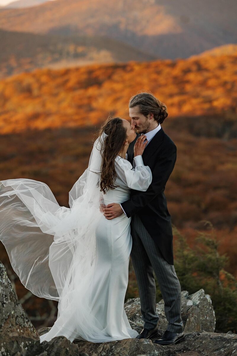 A bride and groom kiss during their sunrise elopement in Shenandoah. There are peak foliage colors surrounding them in the background.