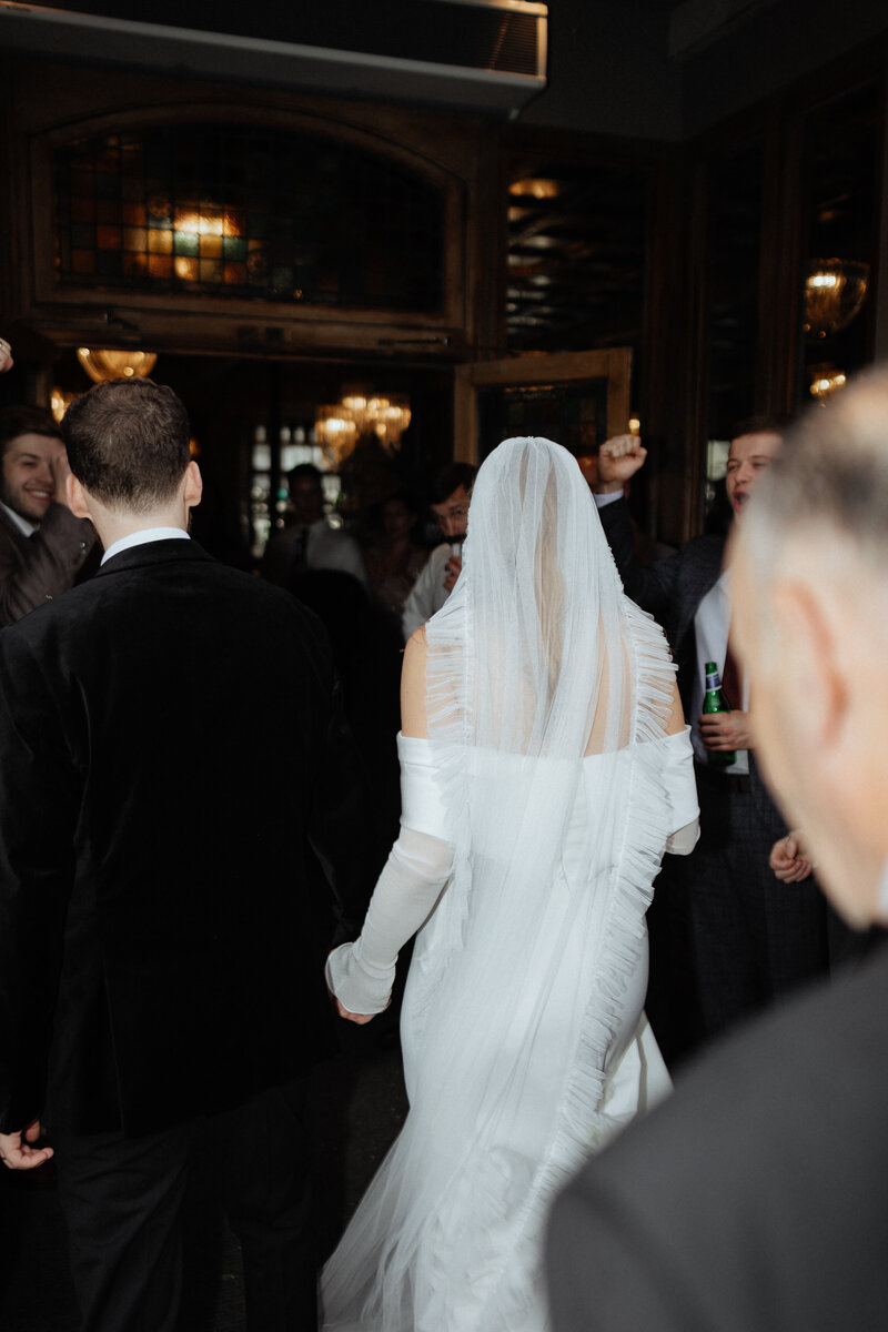 Back view of bride in off-shoulder dress and groom entering an ornate wedding reception venue, photographed in editorial style.