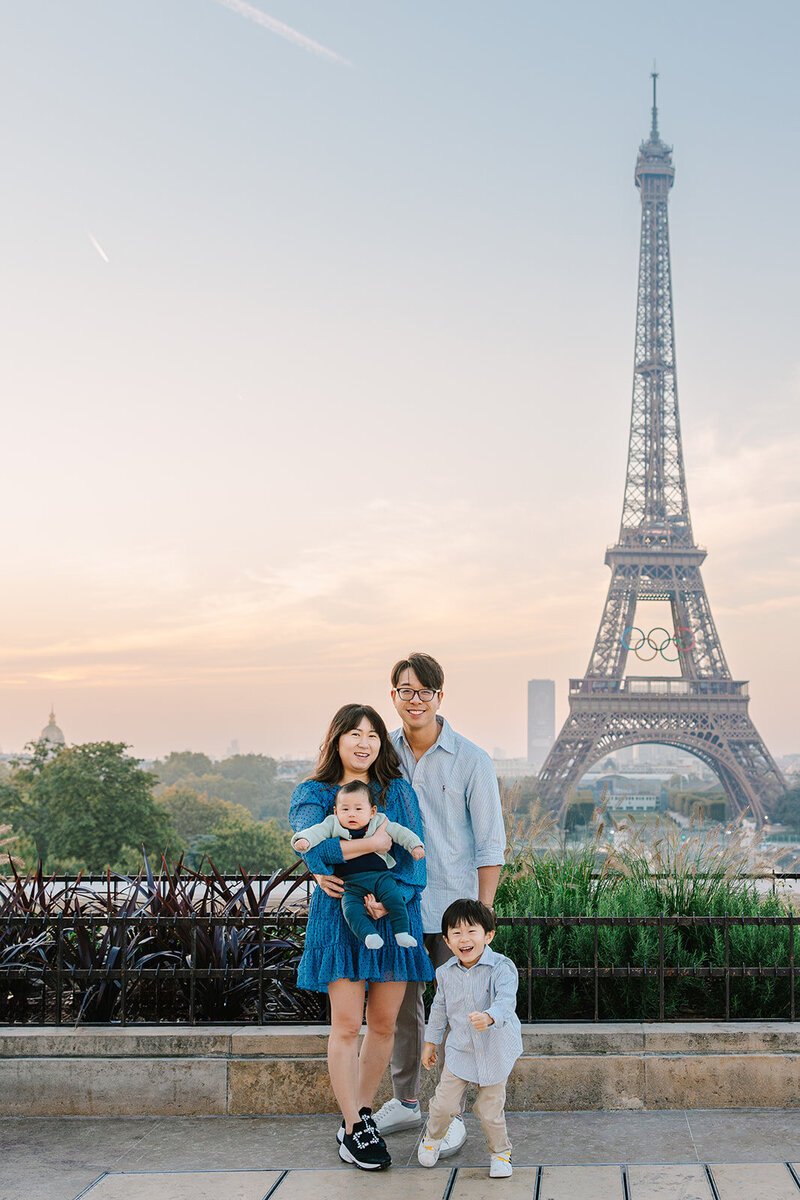 Family of four — mother, father, young son, and baby — smiling and posing by the Eiffel Tower at Trocadéro during a Paris family photoshoot.