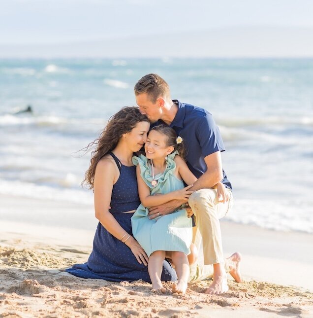 Mom and dad have blue on and daughter has Teal and they're sitting in the sand with the Pacific Ocean in the background.