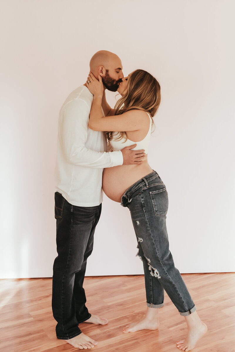 An expecting couple share a kiss in a white studio.