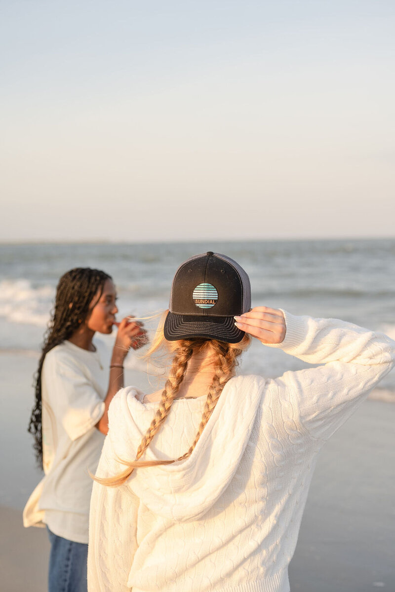 young woman wearing a backwards as her golden braids shine in the sunlight on the beach