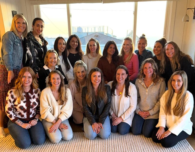 A group of 18 women smiling and sitting together indoors at a Rocketship Mastermind event, symbolizing community, support, and powerful connection among female entrepreneurs. The setting is warm and bright, with a window in the background letting in natural light.