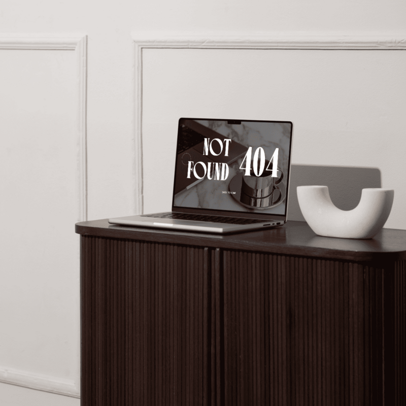 A laptop sits on a dark wood cabinet, displaying a "Not Found 404" error message on its screen. A white, crescent-shaped bowl is on the right side of the laptop. The background features a paneled wall.