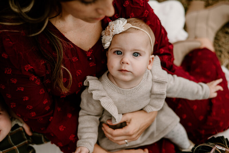 Twin baby girl sitting on moms lap wearing a boy and tan dress while mom is wearing a red dress sitting on a blanket in Gambrills Maryland