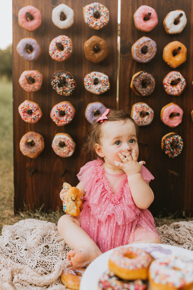 baby girl sitting in front of a donut wall eating a donut