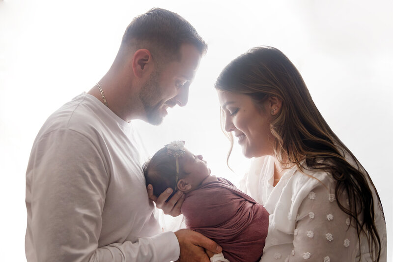 Mom and Dad hold baby together in front of window during photo session.