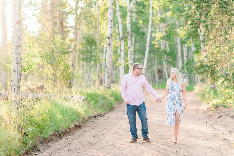 A couple walks hand in hand down a trail lined with green aspen trees in the mountains.