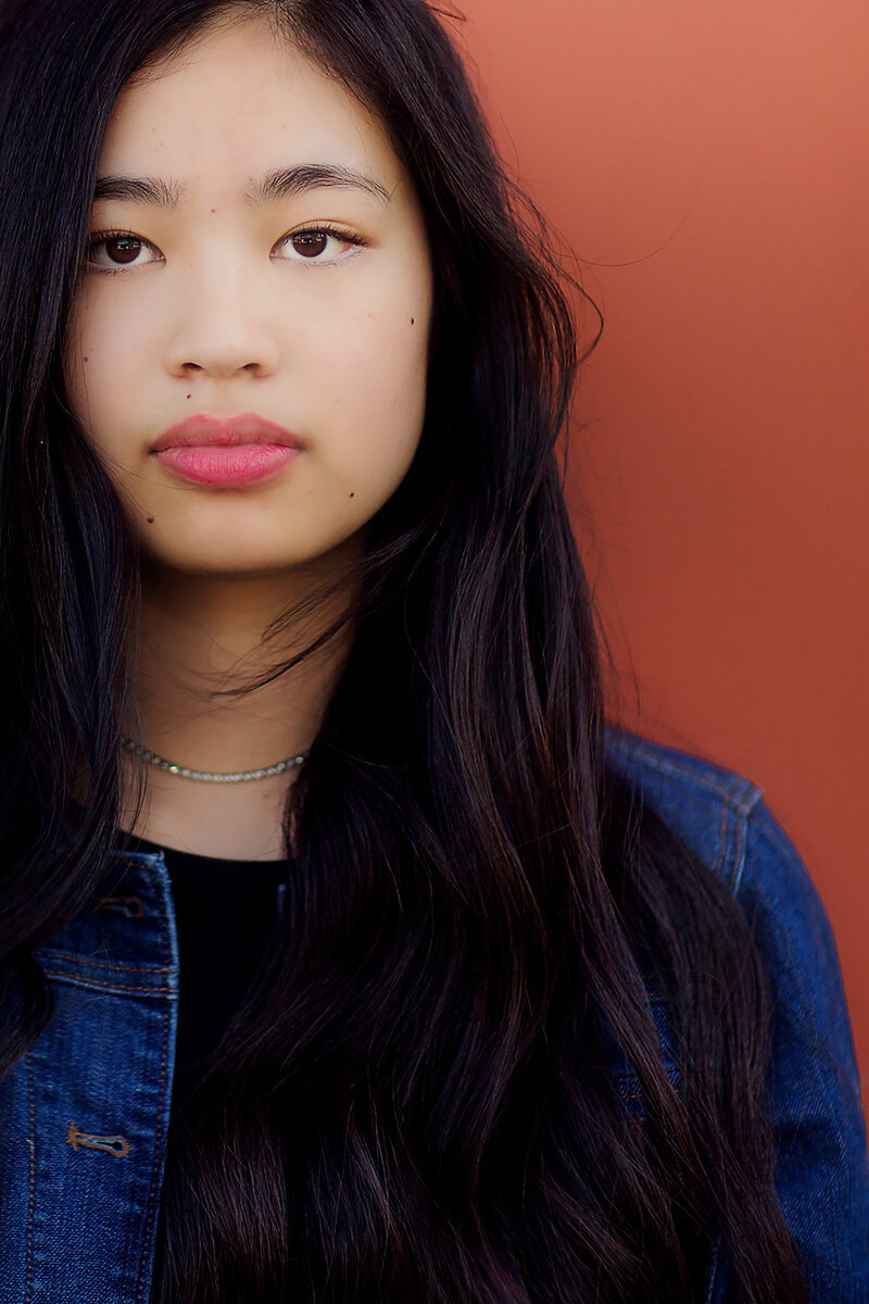 Portrait of a teenage girl with beautiful long, dark hair, looking intensely at the camera. There is a burnt red wall behind her.