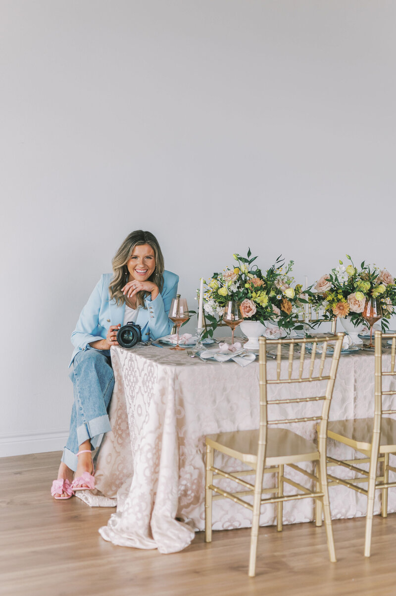 Headshot of Lindsey Kristian in a blue blazer and jeans holding her camera and sitting at a table with her face leaning on her hand