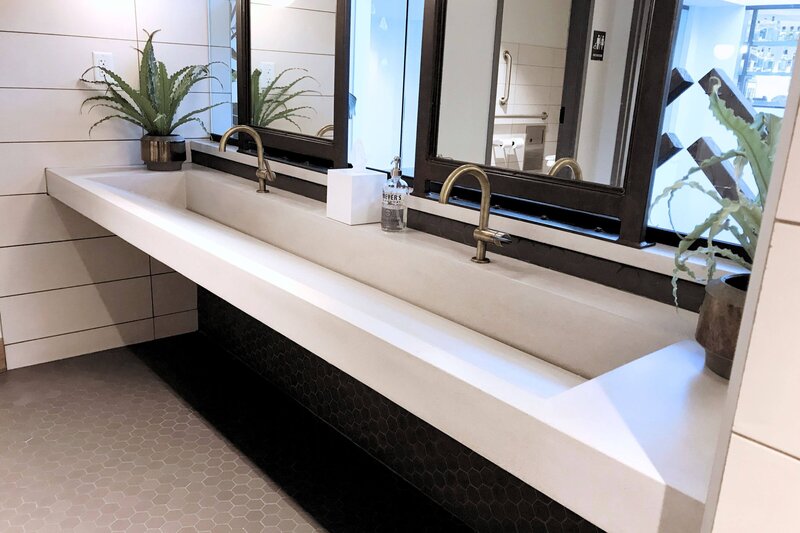 White concrete countertop with integrated ramp sink. Ramp sink is ADA-compliant. Brass faucets and greenery soften the black mirrors. This space was designed for an event center women's bathroom. nter.