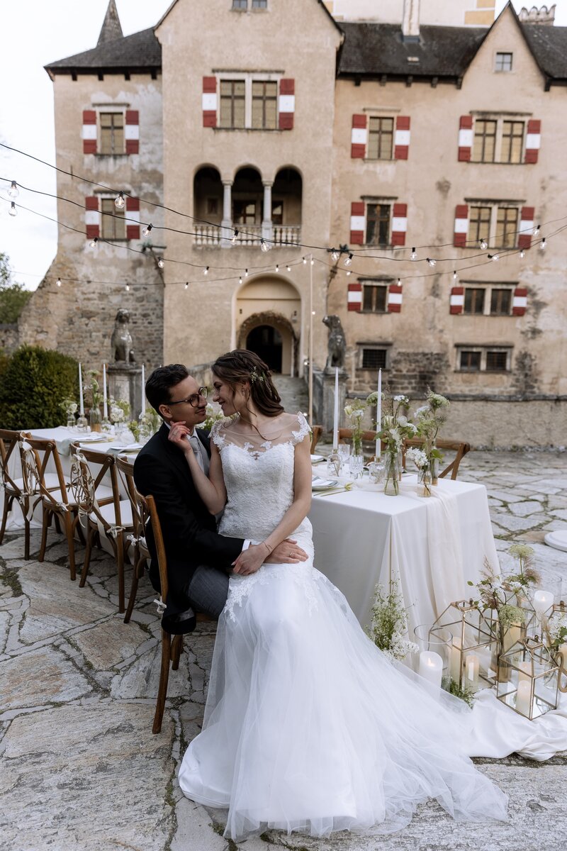Braut sitzt auf Schoss des Braeutigams vor der gedeckten Hochzeitstafel