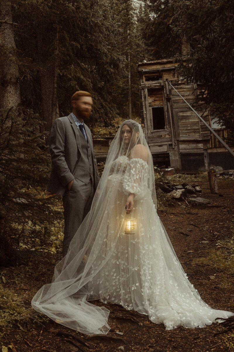 A couple standing in the Colorado woods by an old mine with a lantern dressed in traditional wedding wear.