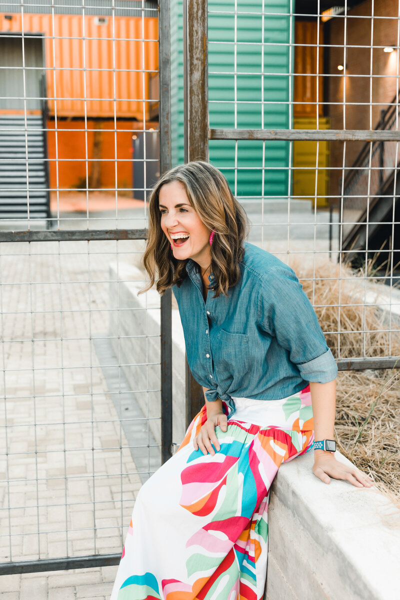 woman wearing denim top and bright skirt laughing in front of multi color background