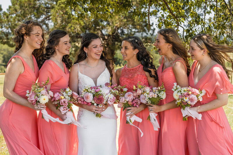 Bride laughs with her bridesmaids while standing in shade under trees.