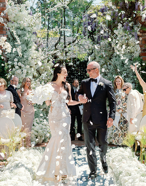 A newly married man and woman are holding hands and smiling, as they walk down an outdoor garden’s aisle surrounded by flowers under an opulent canopy of lights and flowers. Guests, wearing tasteful selections of clothing that reflect a carefully curated color palette, are cheering and clapping as they throw rose petals