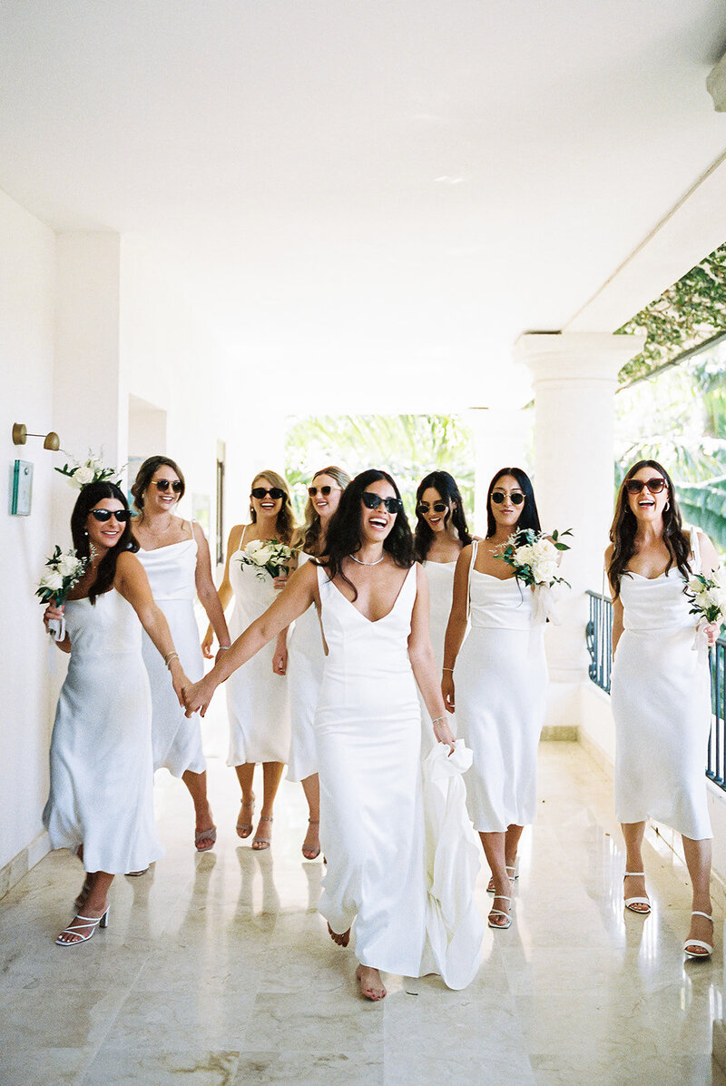 The bride and her bridesmaids in matching white dresses walk through a sunlit corridor at Casa Colonial Hotel, laughing before the ceremony. Captured on film by Asia Pimentel, Caribbean destination wedding photographer.