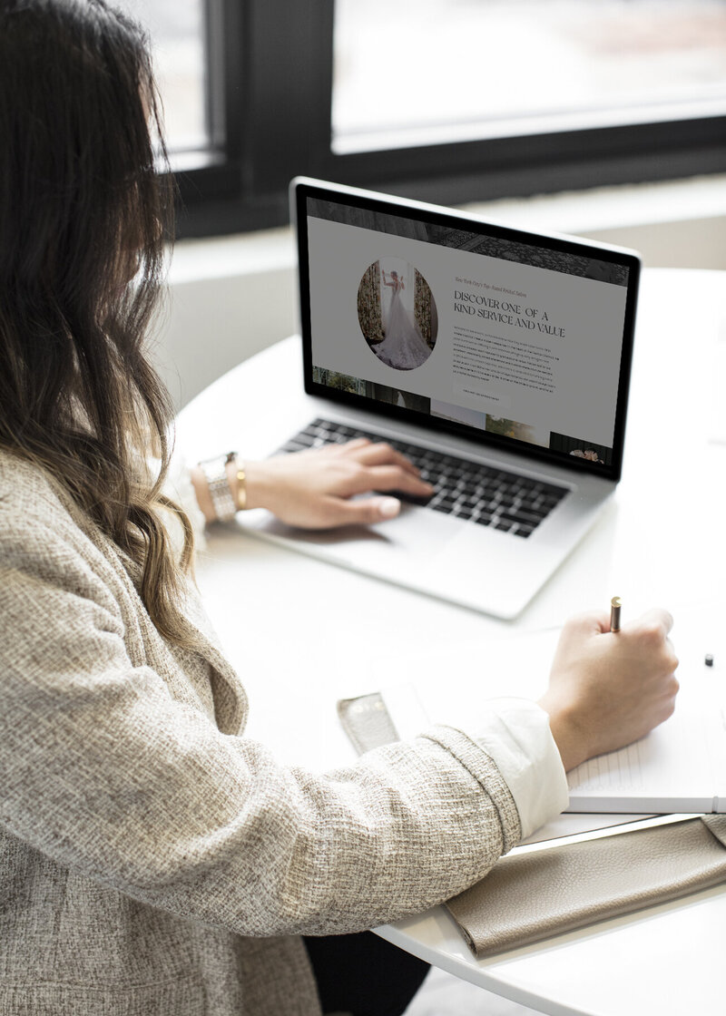 A woman working on a brand website on her laptop at a white desk, taking notes with a gold pen.