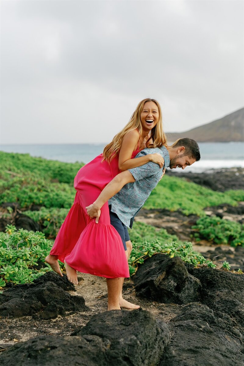 A person on their partner's back as they stand on a rocky path next to the ocean 