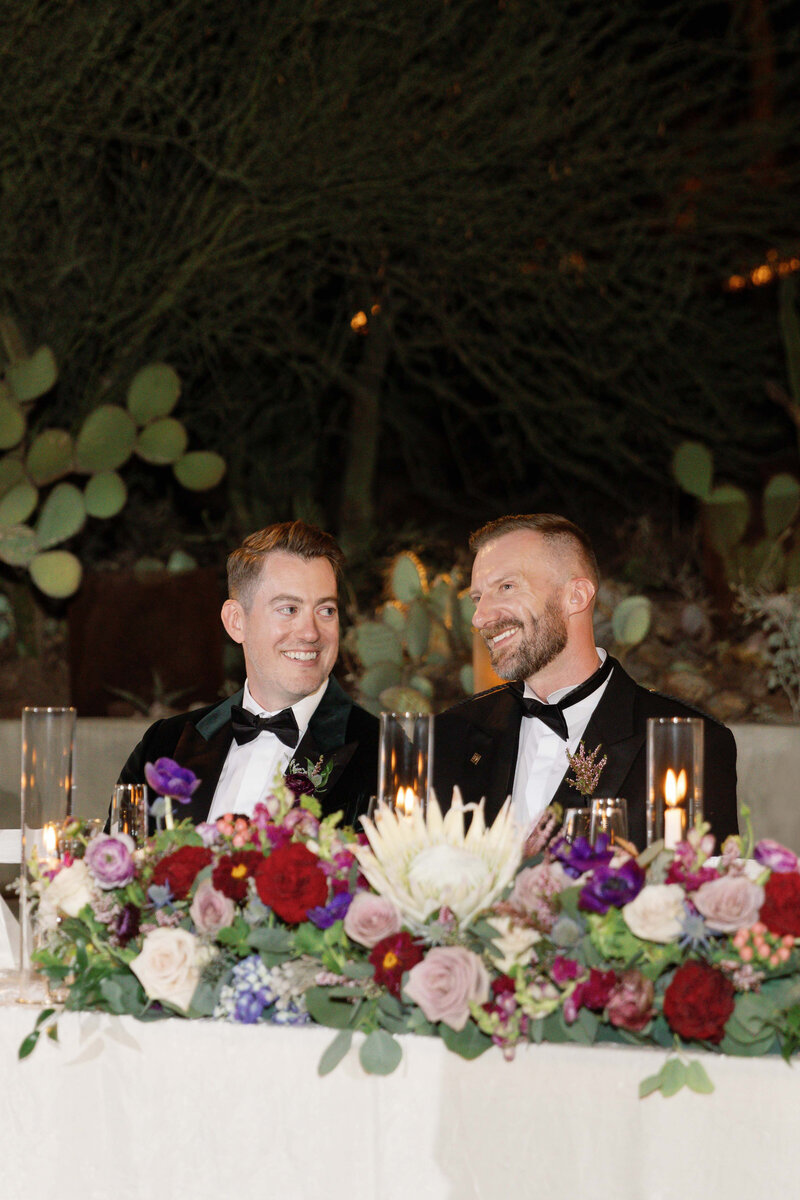 Two grooms sit at sweetheart table during their wedding and smile towards each other.