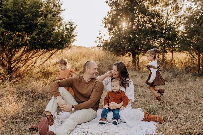 A family of five sits on a blanket for a golden hour photography session. Two sisters play around while mom and dad smile at each other and hold the youngest toddler boy.