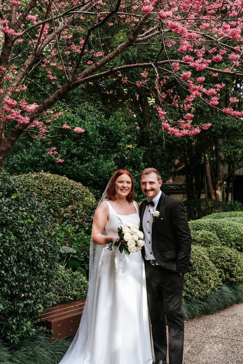 A bride and groom standing in the botanical gardens with trees and flowers in the background