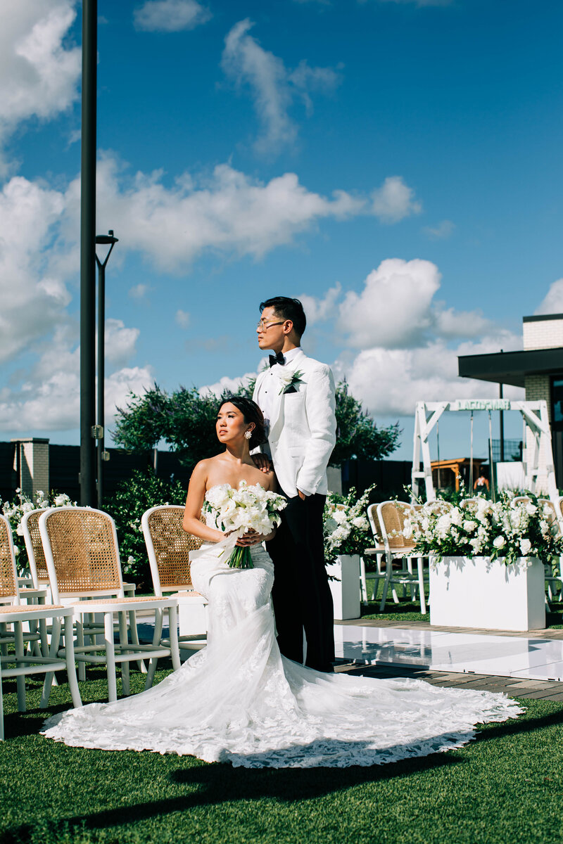 Bride and groom portrait amid rows of white cane-back chairs with white florals under a bright blue Houston sky.