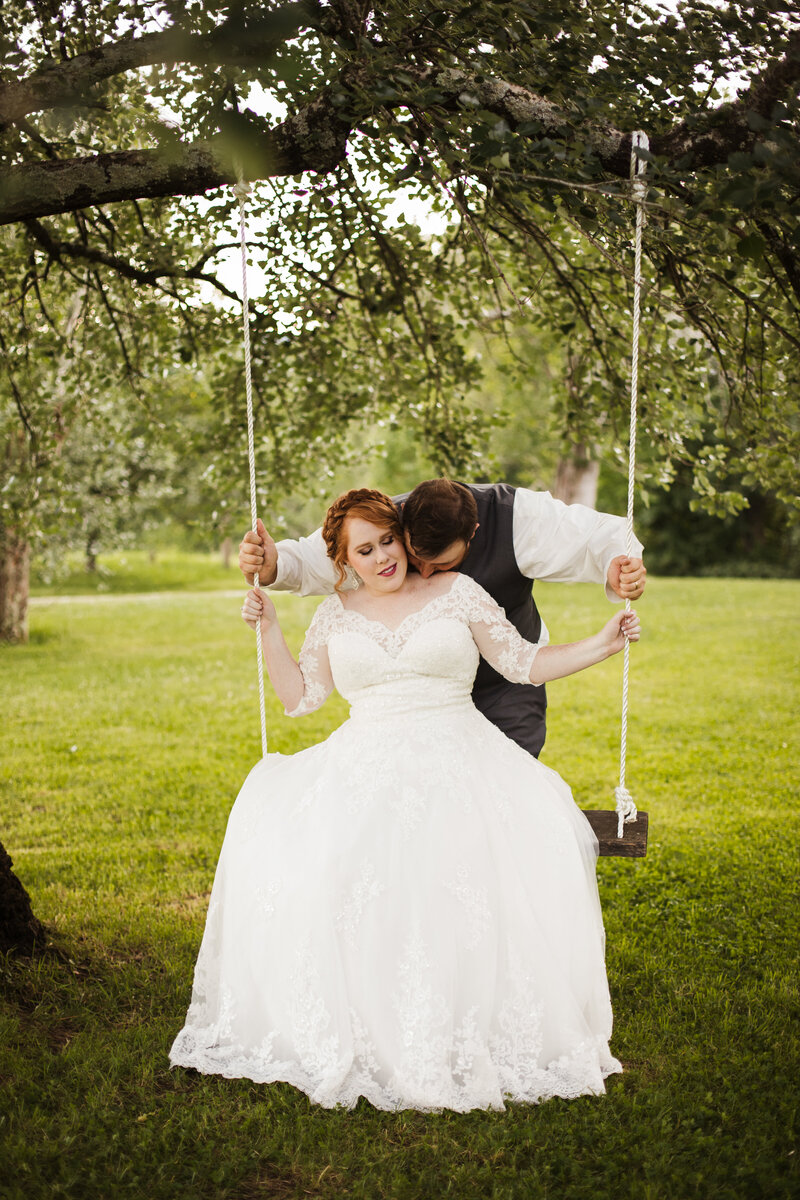 bride sitting on swing while groom kisses her shoulder