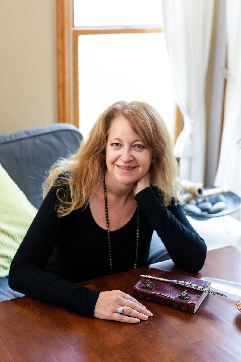Cyndi Stewart sits at a wooden table with a journal and pen, smiling and relaxed in a softly lit room with natural light.