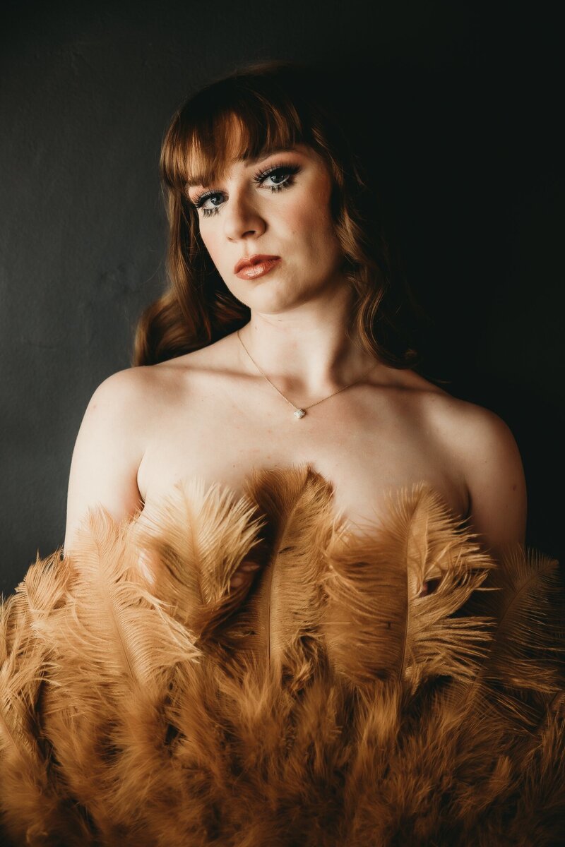 Women holds feather fan in front of her chest looking directly into the camera
