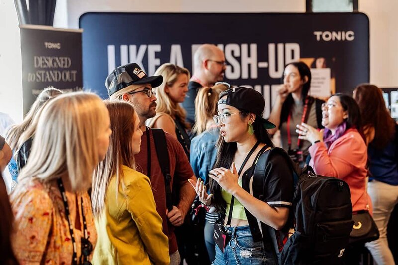 Designers and creatives engage in conversation at a packed event, standing in front of a booth with TONIC branding and the phrase ‘LIKE A PUSH-UP BRA’ partially visible on a backdrop.