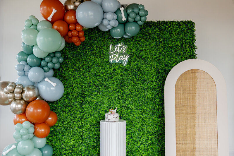 Boxwood greenery wall with rattan arch, dog balloon garland and a cake pedestal.