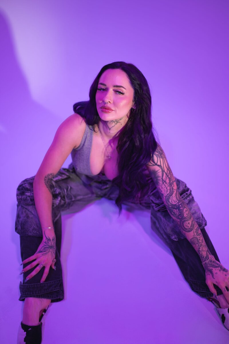 Woman with long, dark hair poses for the camera while sitting on the floor in front of a purple backdrop.