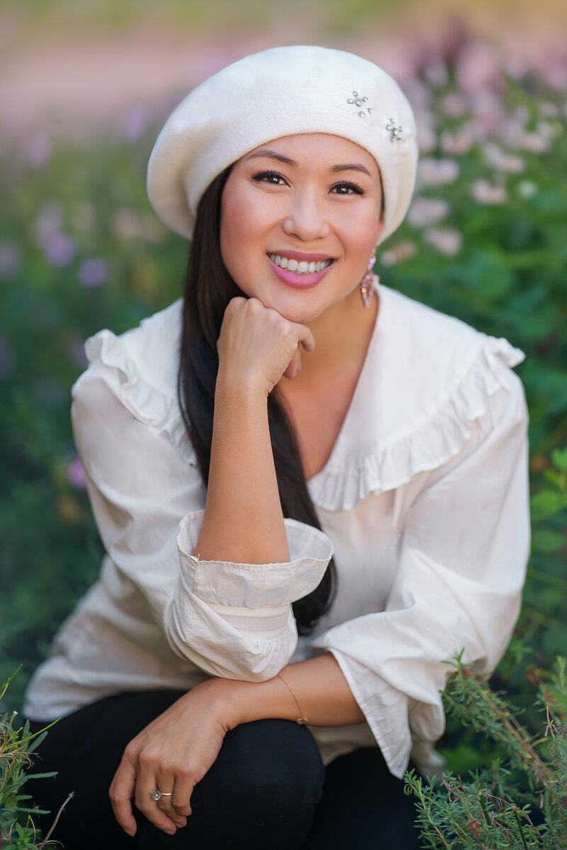 Headshot of woman posing with her fist on her chin wearing a white beret with crystals and a white button up shirt with a Peter Pan collar sitting in front of a field of flowers