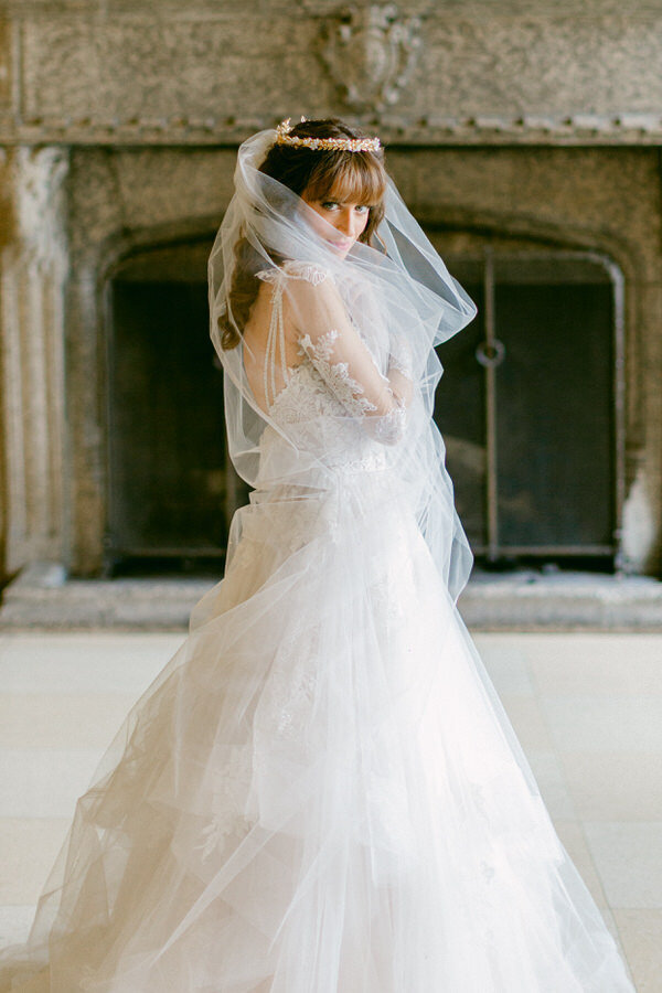 This captivating portrait captures a bride standing gracefully at the Banff Springs Hotel, looking into the camera. Behind her, a grand fireplace adds warmth to the scene, while large windows on her right bathe the room in natural light, highlighting her elegance.